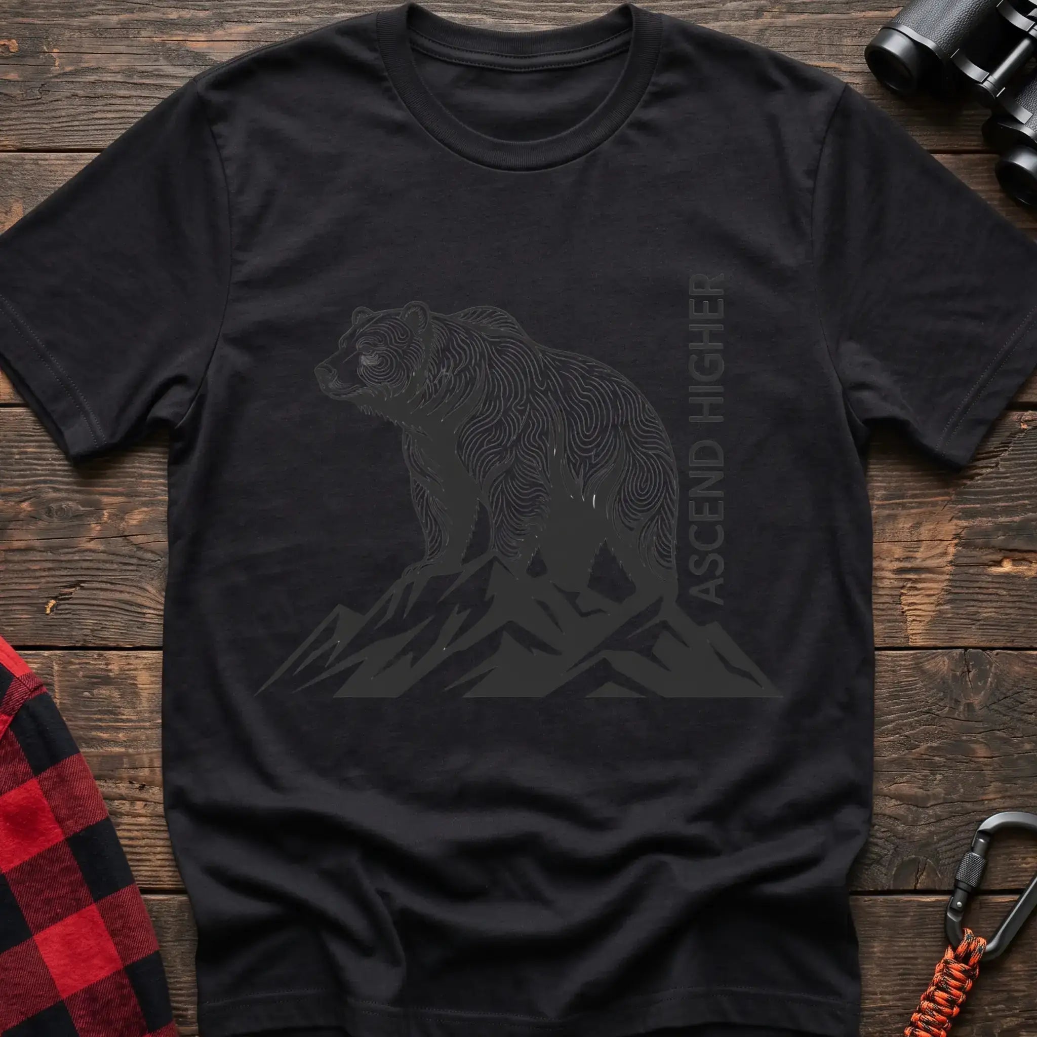Black t-shirt with a black illustration of a bear standing on mountain peaks, with the vertical text “ASCEND HIGHER” beside it, laid flat on a wooden surface.