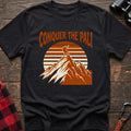 Black t-shirt with orange text reading Conquer the Pali above a hiker standing on a mountain peak with a retro sun graphic