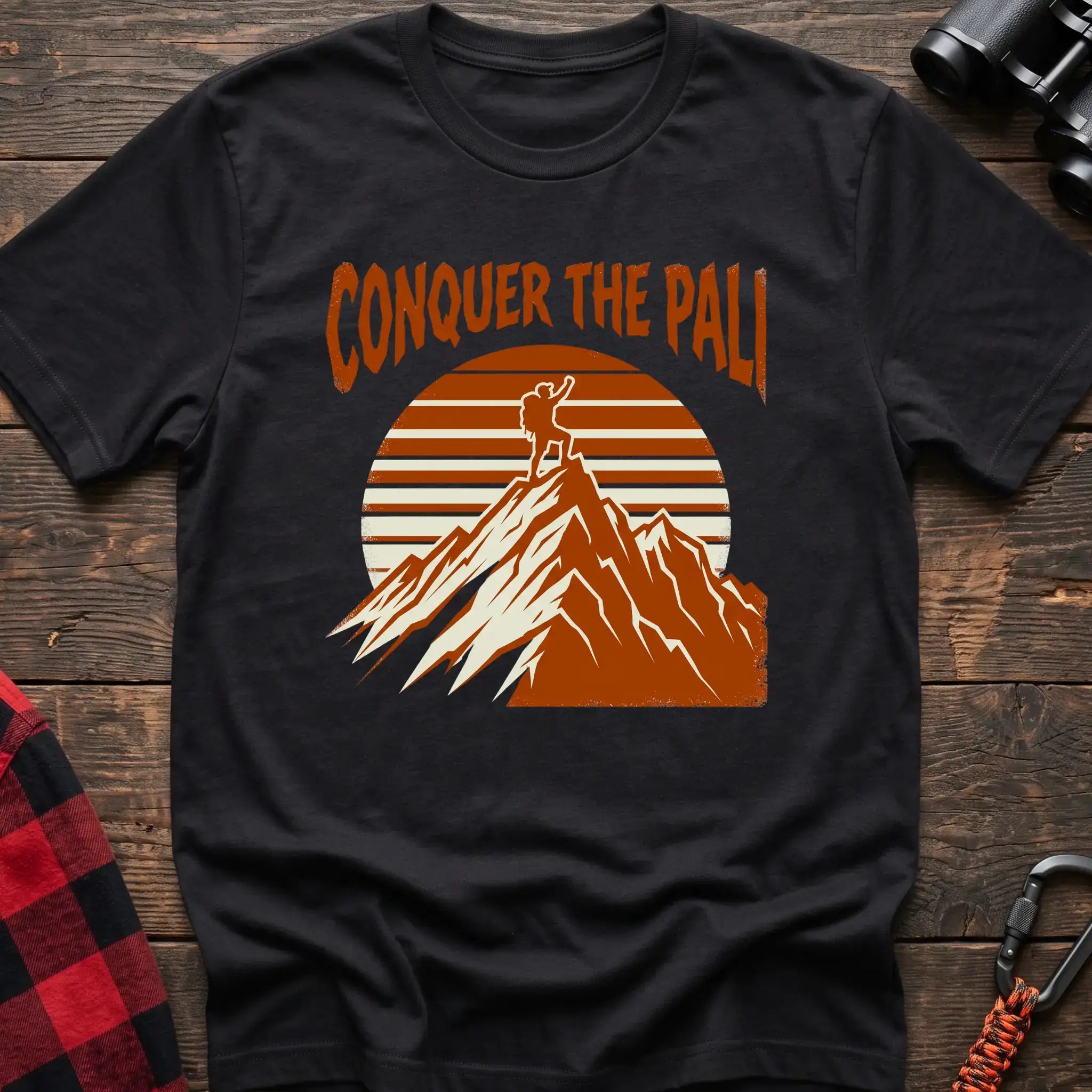 Black t-shirt with orange text reading Conquer the Pali above a hiker standing on a mountain peak with a retro sun graphic