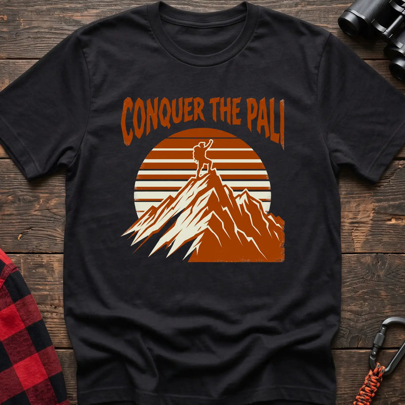 Black t-shirt with orange text reading Conquer the Pali above a hiker standing on a mountain peak with a retro sun graphic