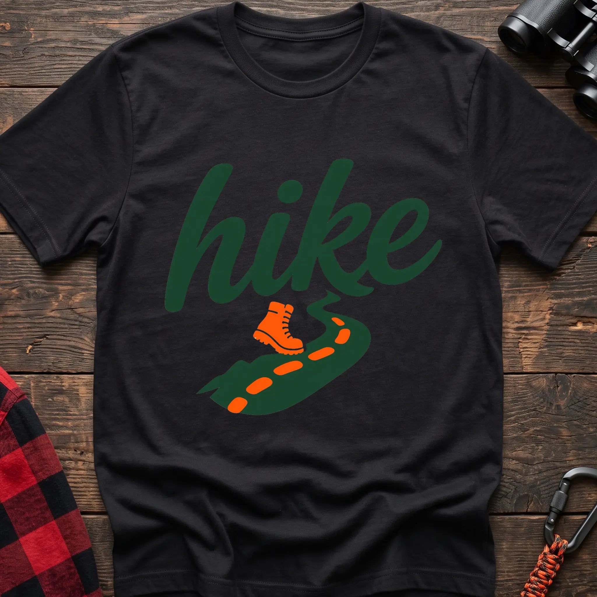 Black cotton t-shirt with green cursive hike script, orange hiking boot, and footprint trail graphic, flat lay on rustic wood with hiking gear