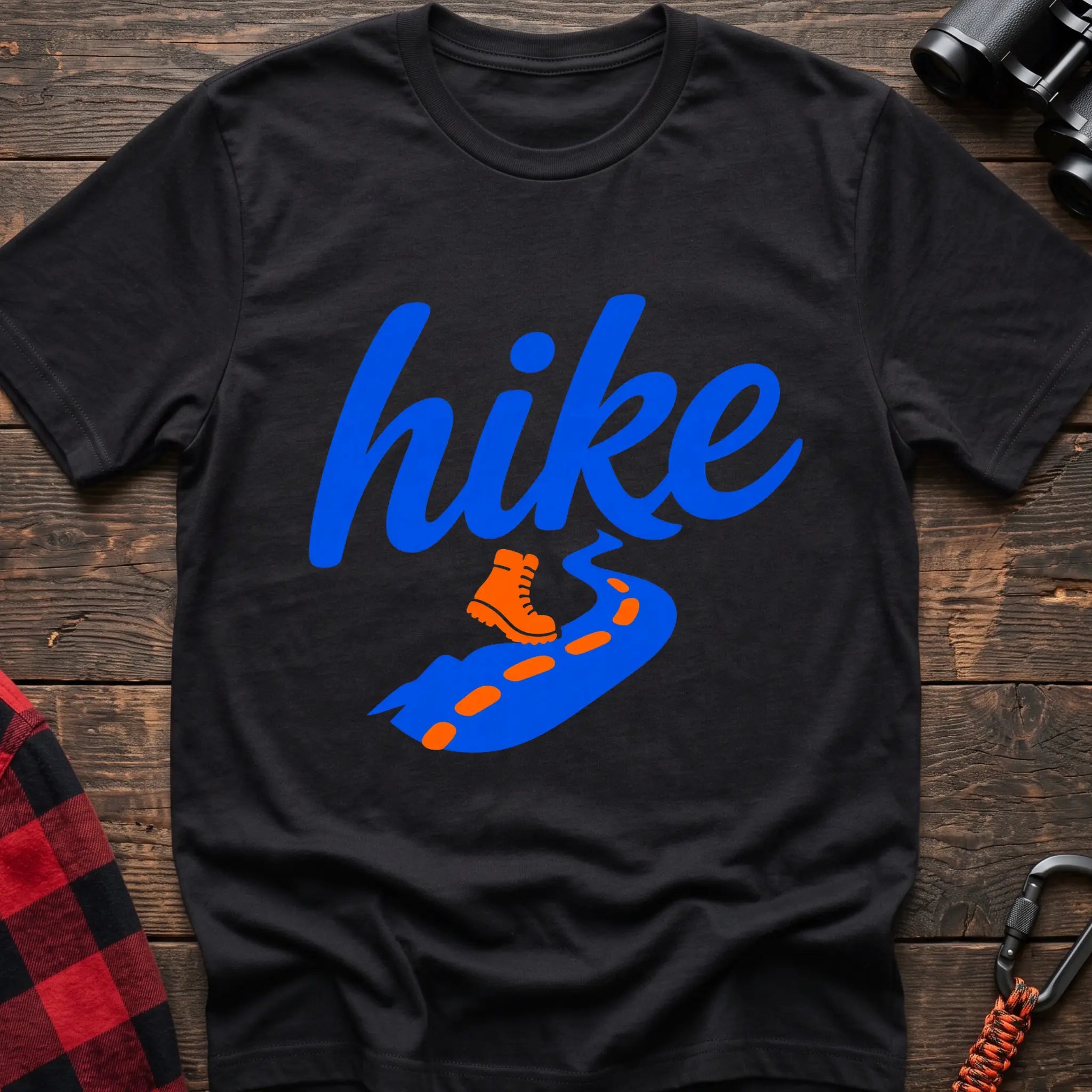 Black cotton t-shirt with royal blue cursive hike script, orange hiking boot, and blue trail with orange footprints, flat lay on rustic wood with hiking gear