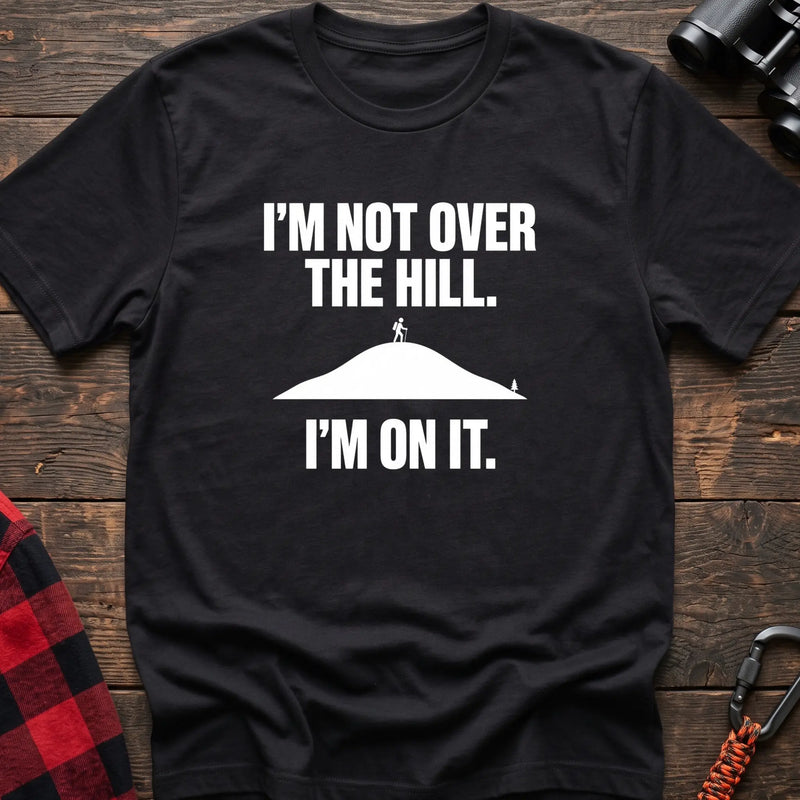 Black cotton t-shirt with a hiker on a hill graphic and I’m Not Over the Hill I’m On It text on rustic wood