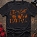 Black t-shirt with bold orange handwritten-style text reading “I THOUGHT THIS WAS A FLAT TRAIL,” laid flat on a wooden surface.