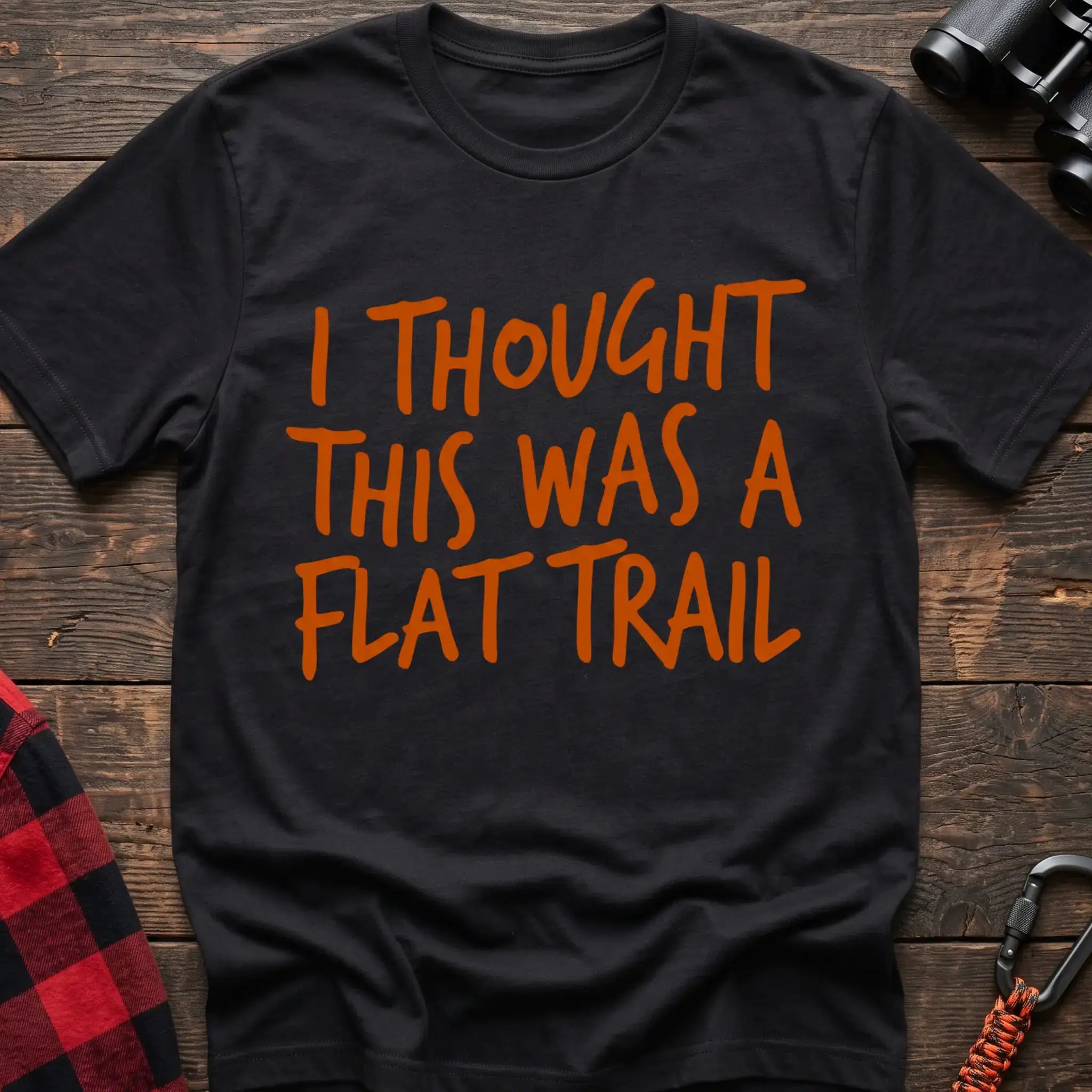 Black t-shirt with bold orange handwritten-style text reading “I THOUGHT THIS WAS A FLAT TRAIL,” laid flat on a wooden surface.