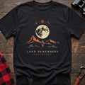 Black cotton t-shirt with a desert hiker and full moon graphic and Land Remembers text on rustic wood
