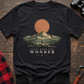 Black cotton t-shirt with a hiker and mountain sunset graphic and Leave Room for Wonder text on rustic wood