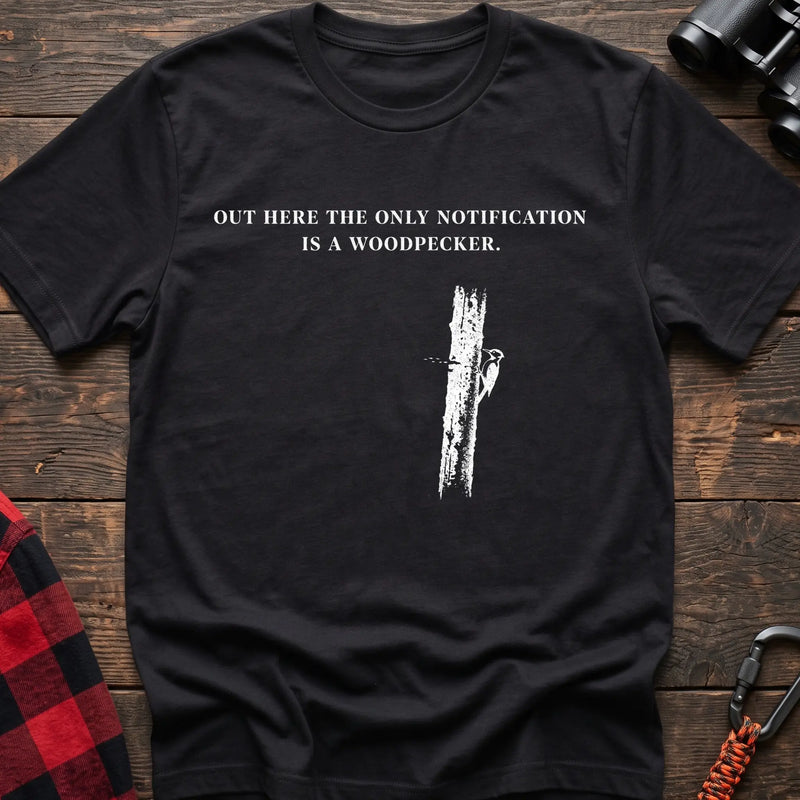 Black cotton t-shirt with a woodpecker graphic and Out Here the Only Notification Is a Woodpecker text on rustic wood