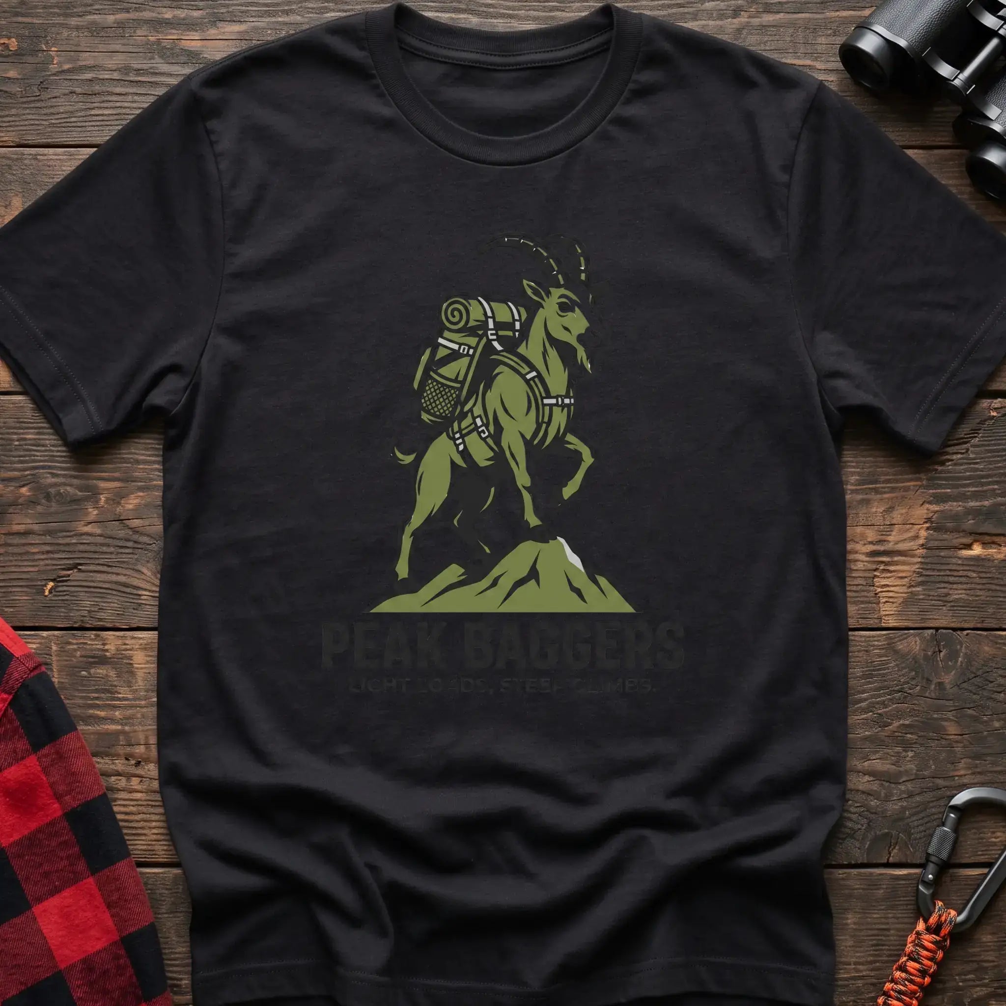 Black t-shirt with an illustration of a mountain goat wearing a backpack standing on a peak, with the text “PEAK BAGGERS” and “LIGHT LOADS, STEEP CLIMBS,” laid flat on a wooden surface.