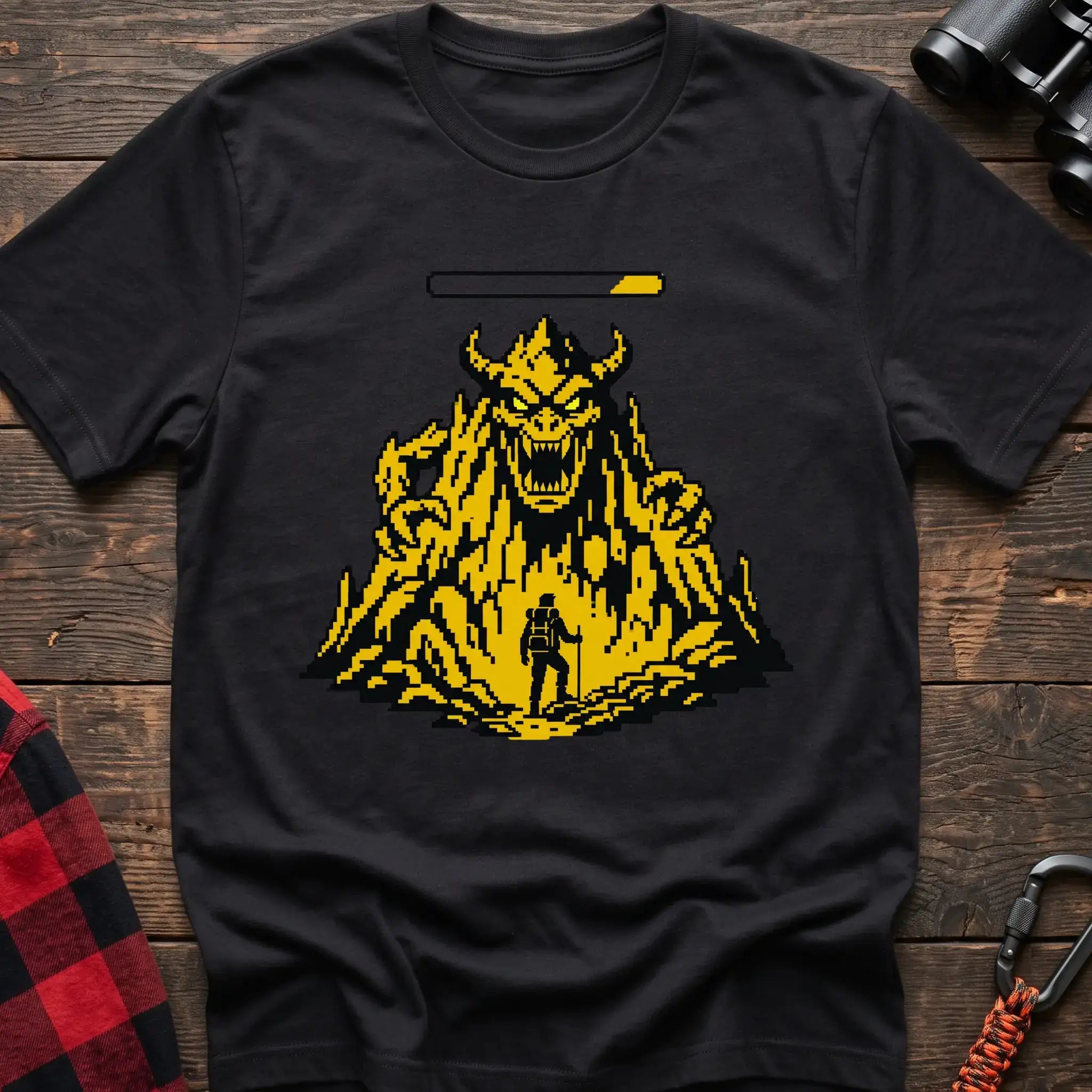 Black t-shirt with a pixel-art illustration of a hiker facing a large horned mountain monster with a health bar above it, laid flat on a wooden surface.