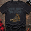 Black t-shirt with an illustration of a dirty hiking boot with rising steam lines, and the text “SMELL THAT? THAT’S VICTORY. (And slightly fermented trail mix),” laid flat on a wooden surface