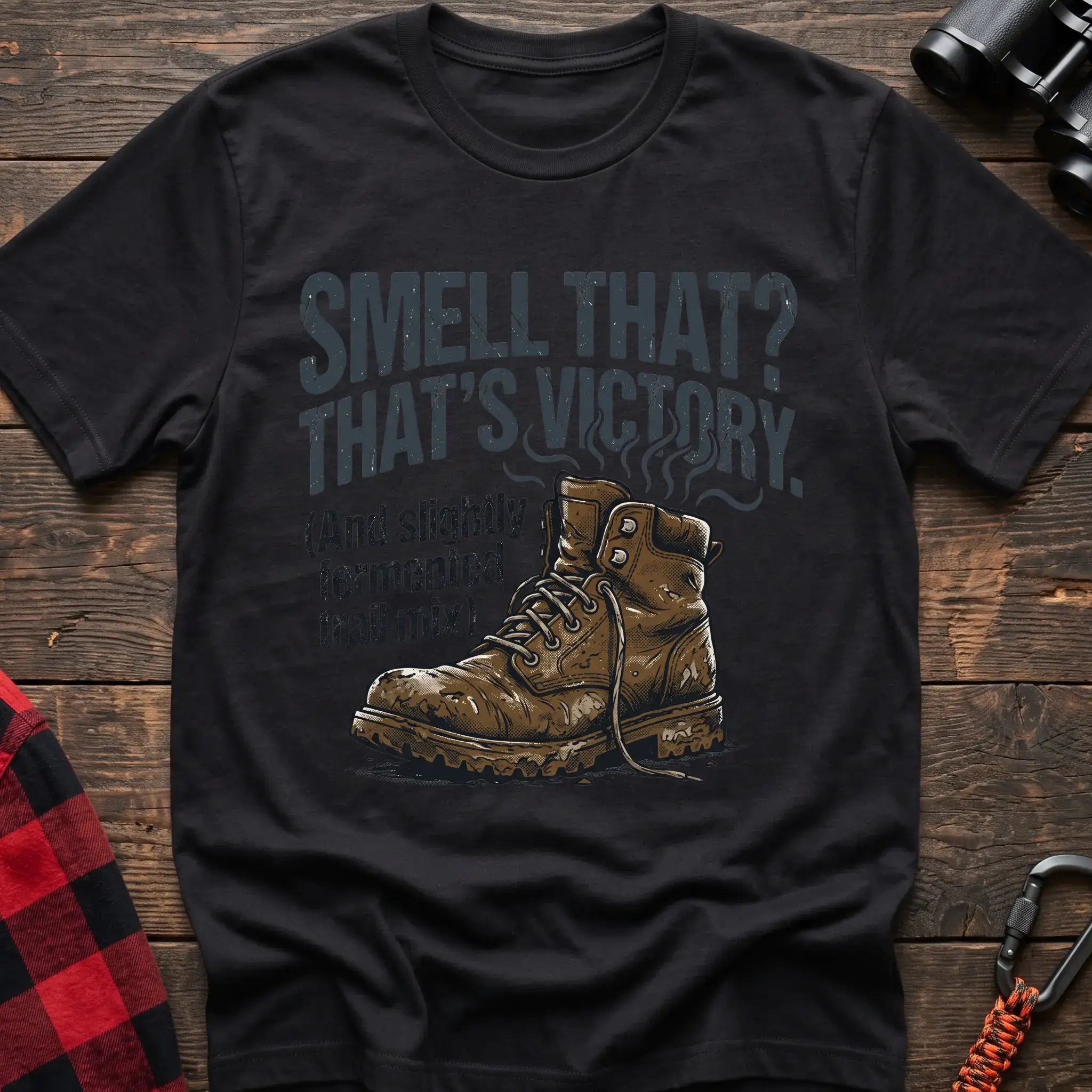Black t-shirt with an illustration of a dirty hiking boot with rising steam lines, and the text “SMELL THAT? THAT’S VICTORY. (And slightly fermented trail mix),” laid flat on a wooden surface