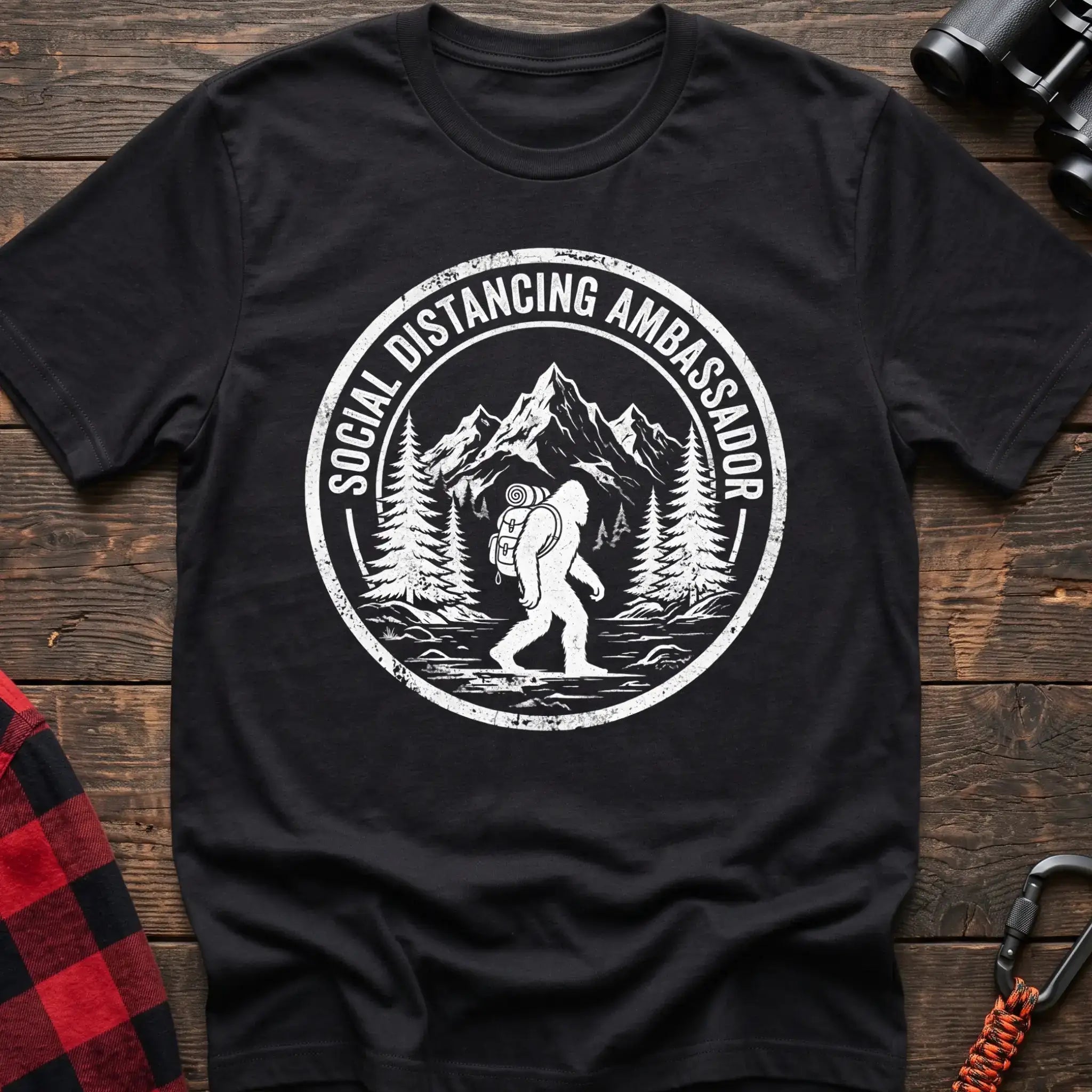 Black t-shirt with white circular badge graphic of Bigfoot hiking with a backpack through mountains and the text Social Distancing Ambassador