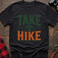 Black t-shirt with distressed green and orange text reading “TAKE A HIKE” with a small mountain graphic between the words, laid flat on a wooden surface.