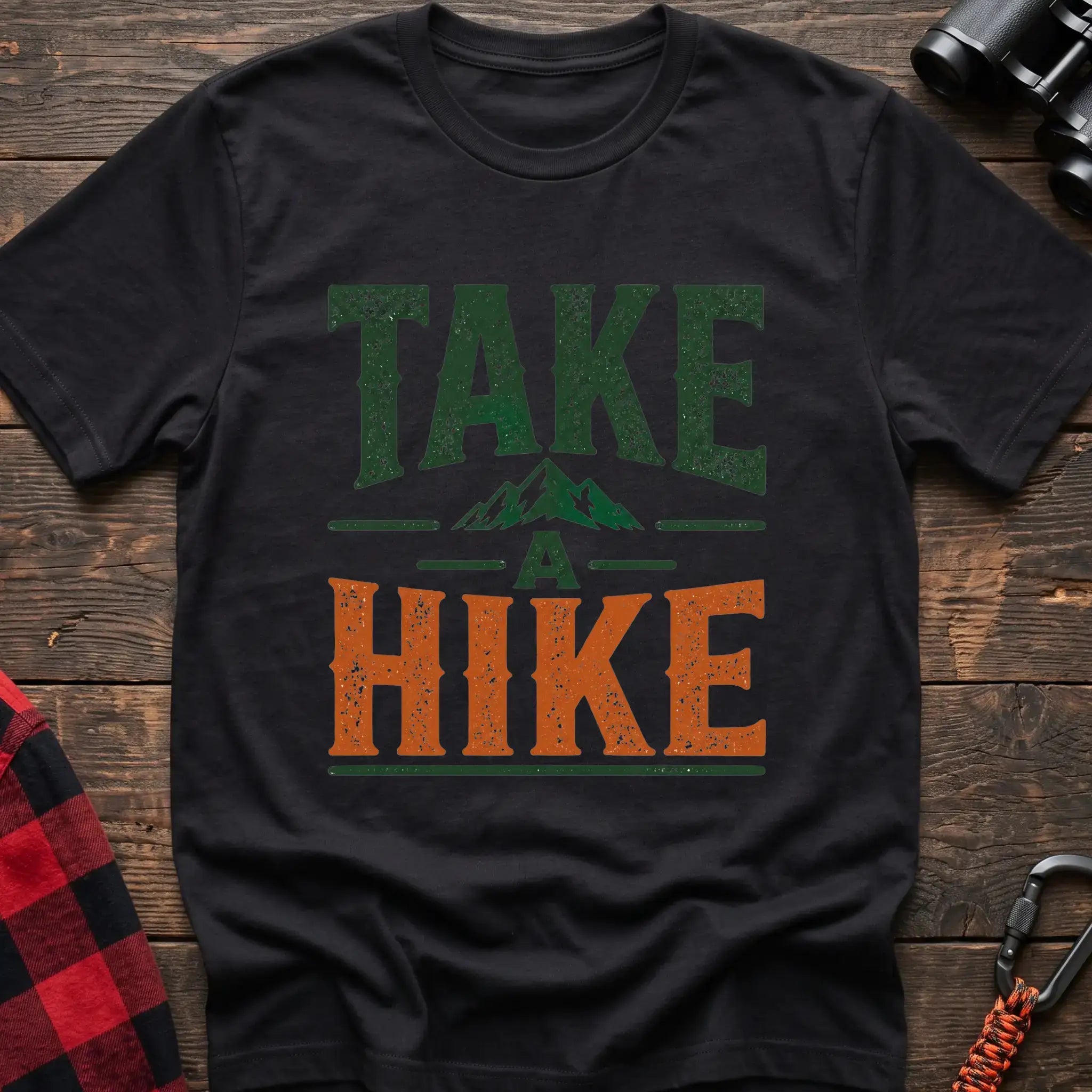 Black t-shirt with distressed green and orange text reading “TAKE A HIKE” with a small mountain graphic between the words, laid flat on a wooden surface.