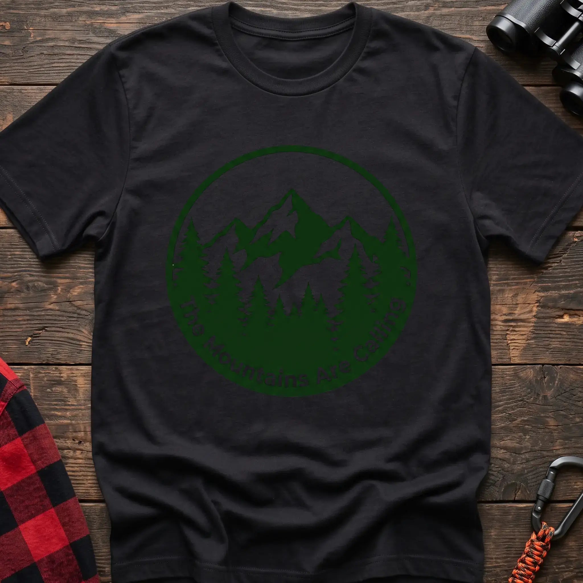 Black t-shirt with a circular dark green design featuring mountain peaks and pine trees, with the text “The Mountains Are Calling,” laid flat on a wooden surface.