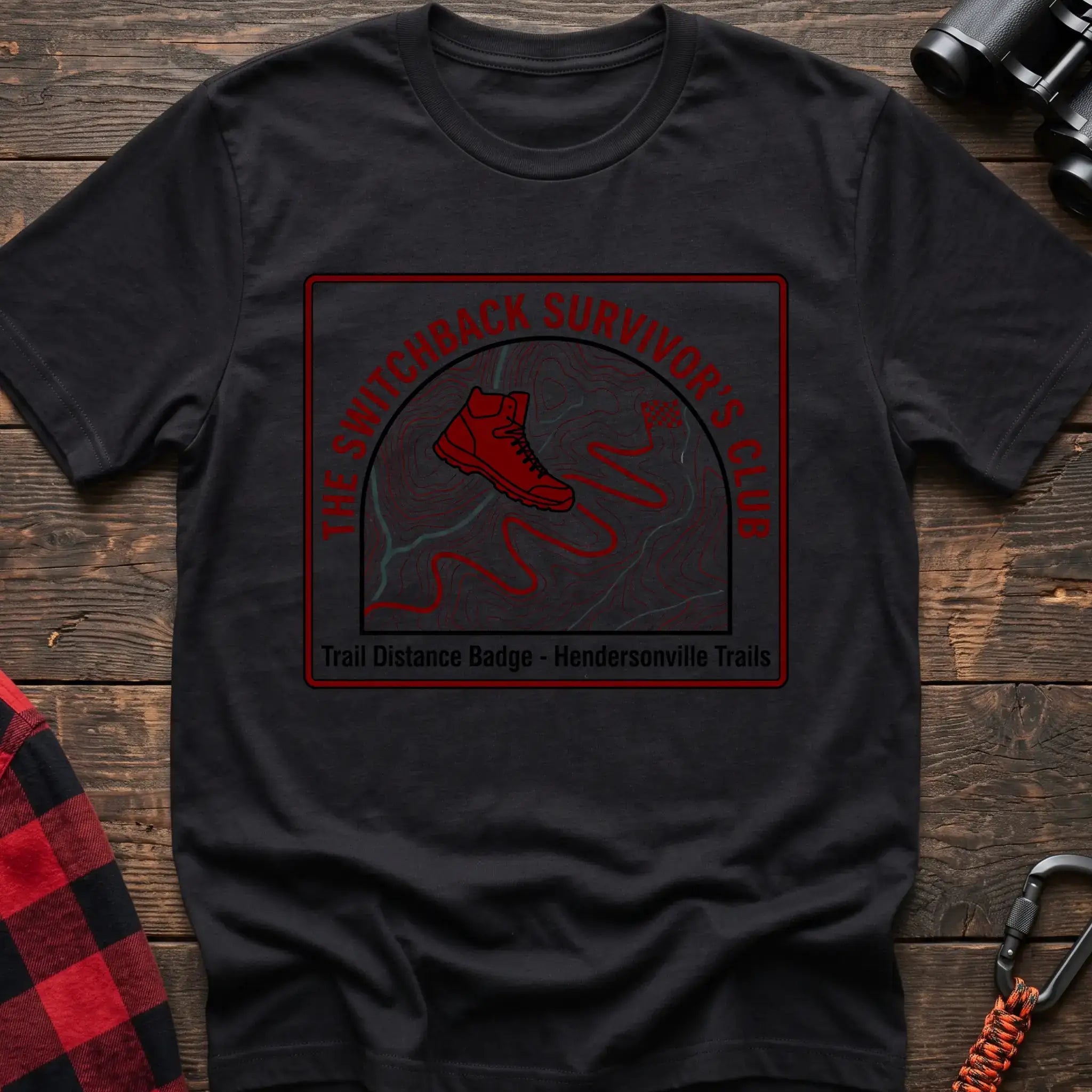 Black t-shirt with a square badge design featuring a red hiking boot on a topographic map with a winding trail, and the text “THE SWITCHBACK SURVIVOR’S CLUB” and “Trail Distance Badge - Hendersonville Trails,” laid flat on a wooden surface.