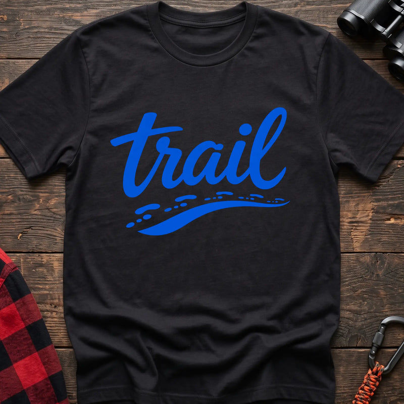 Black cotton t-shirt with blue cursive trail script and dotted path graphic, flat lay on rustic wood with hiking gear