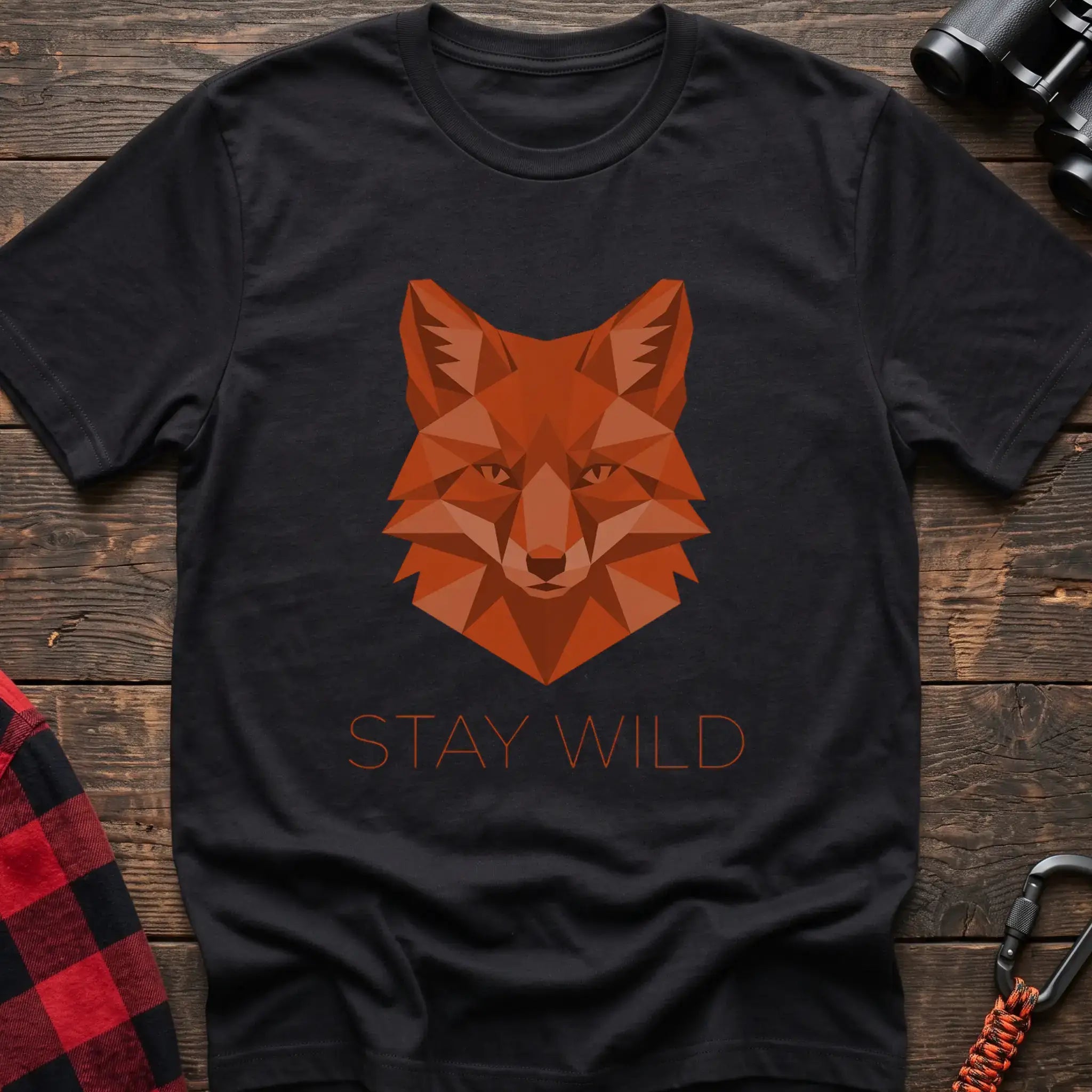 Black t-shirt with a geometric low-poly orange fox head and the text “STAY WILD” printed below in matching orange, laid flat on a wooden surface.
