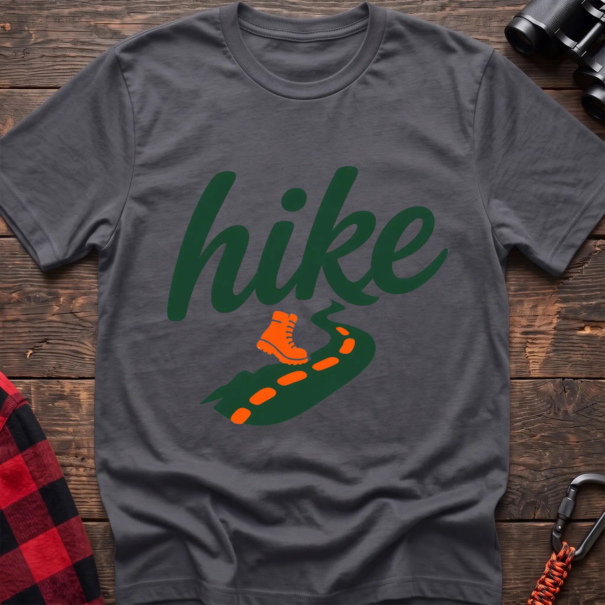 Charcoal cotton t-shirt with green cursive hike script, orange hiking boot, and footprint trail graphic, flat lay on rustic wood with hiking gear
