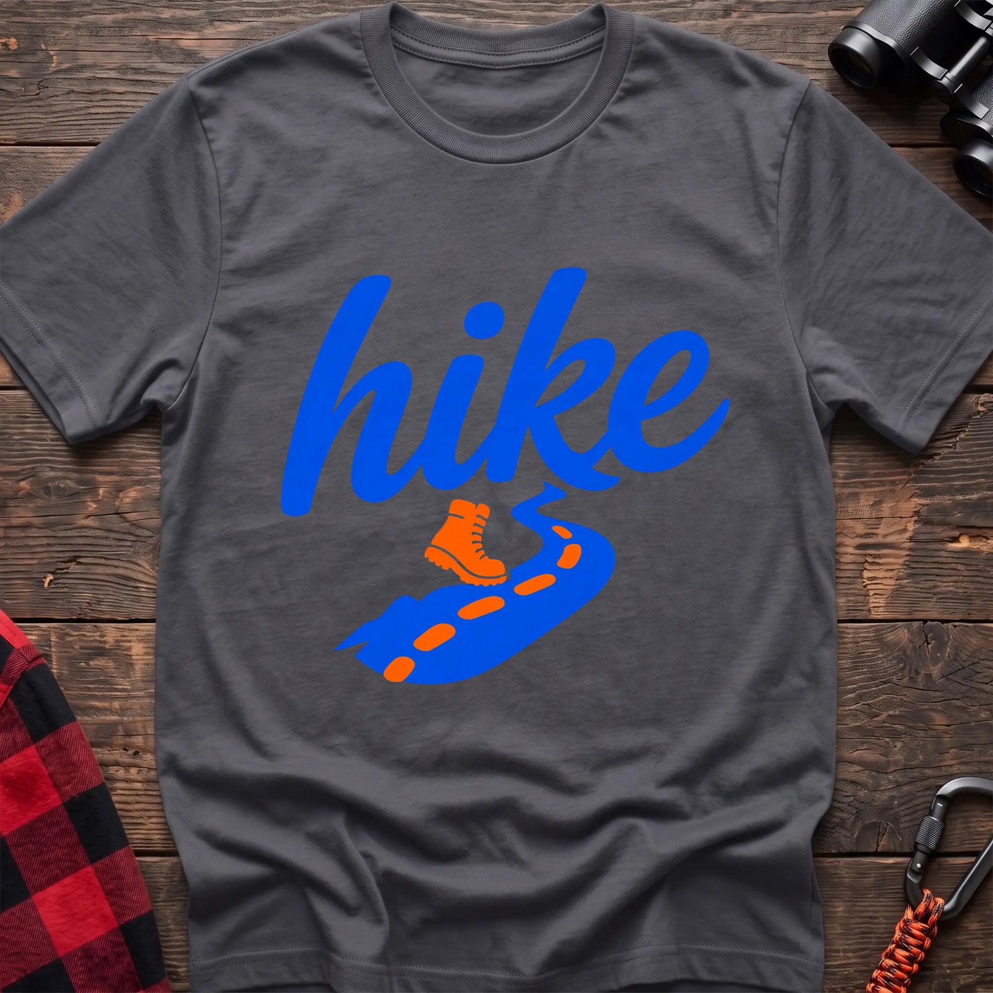 Charcoal cotton t-shirt with royal blue cursive hike script, orange hiking boot, and blue trail with orange footprints, flat lay on rustic wood with hiking gear