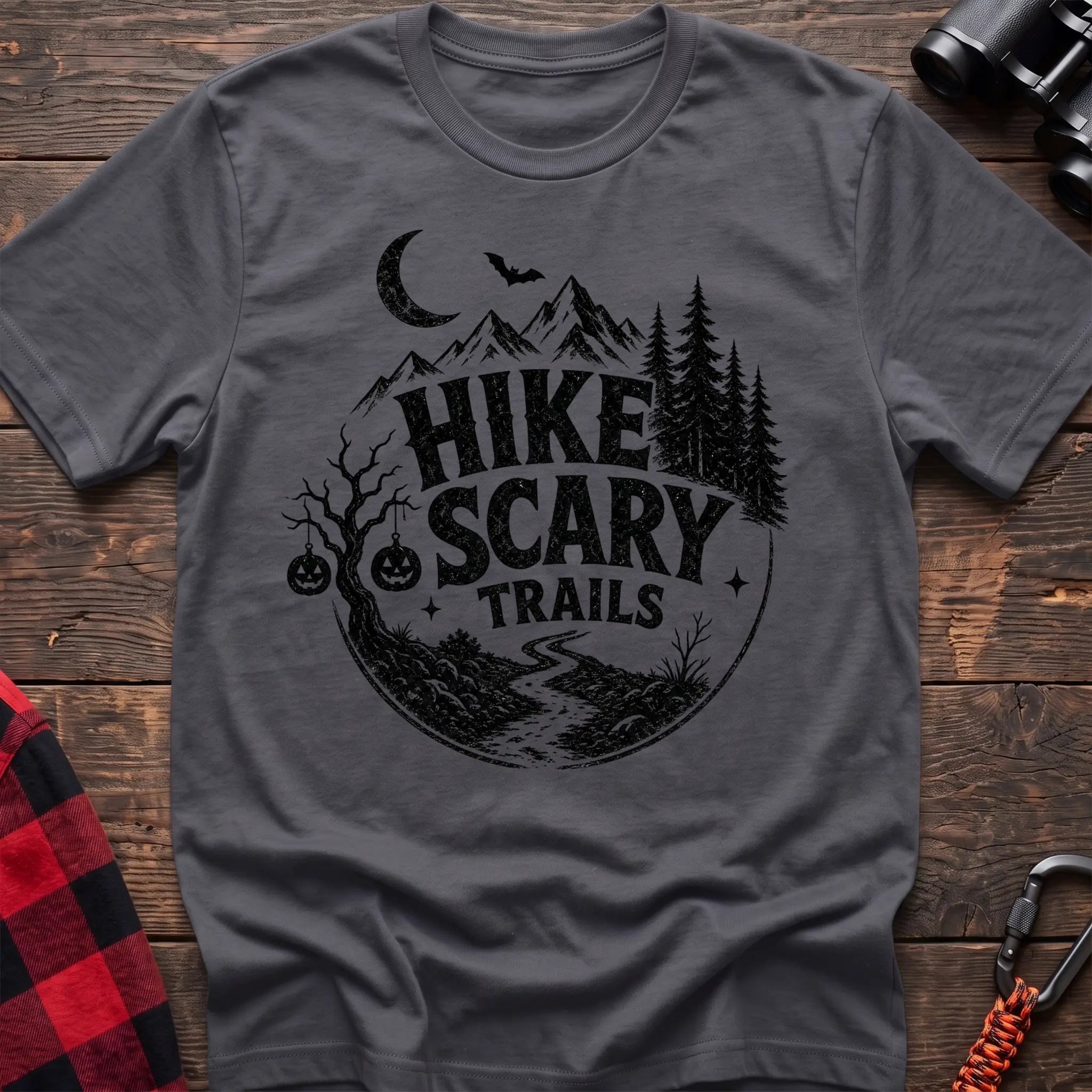 Charcoal cotton t-shirt with vintage Hike Scary Trails graphic of mountains, a crescent moon, bat, jack-o-lanterns, and a winding trail, flat lay on rustic wood with hiking gear