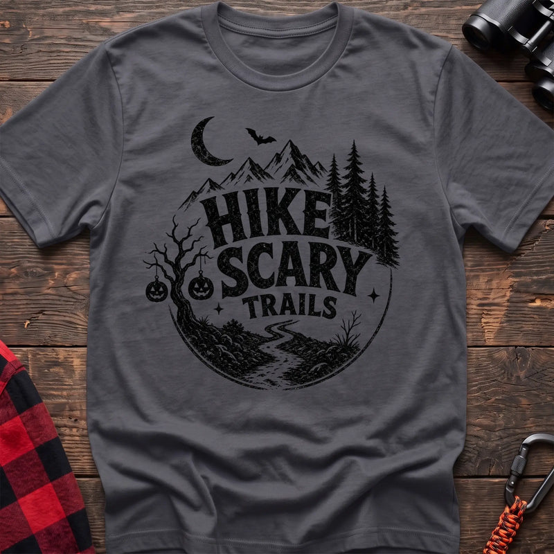 Charcoal cotton t-shirt with vintage Hike Scary Trails graphic of mountains, a crescent moon, bat, jack-o-lanterns, and a winding trail, flat lay on rustic wood with hiking gear