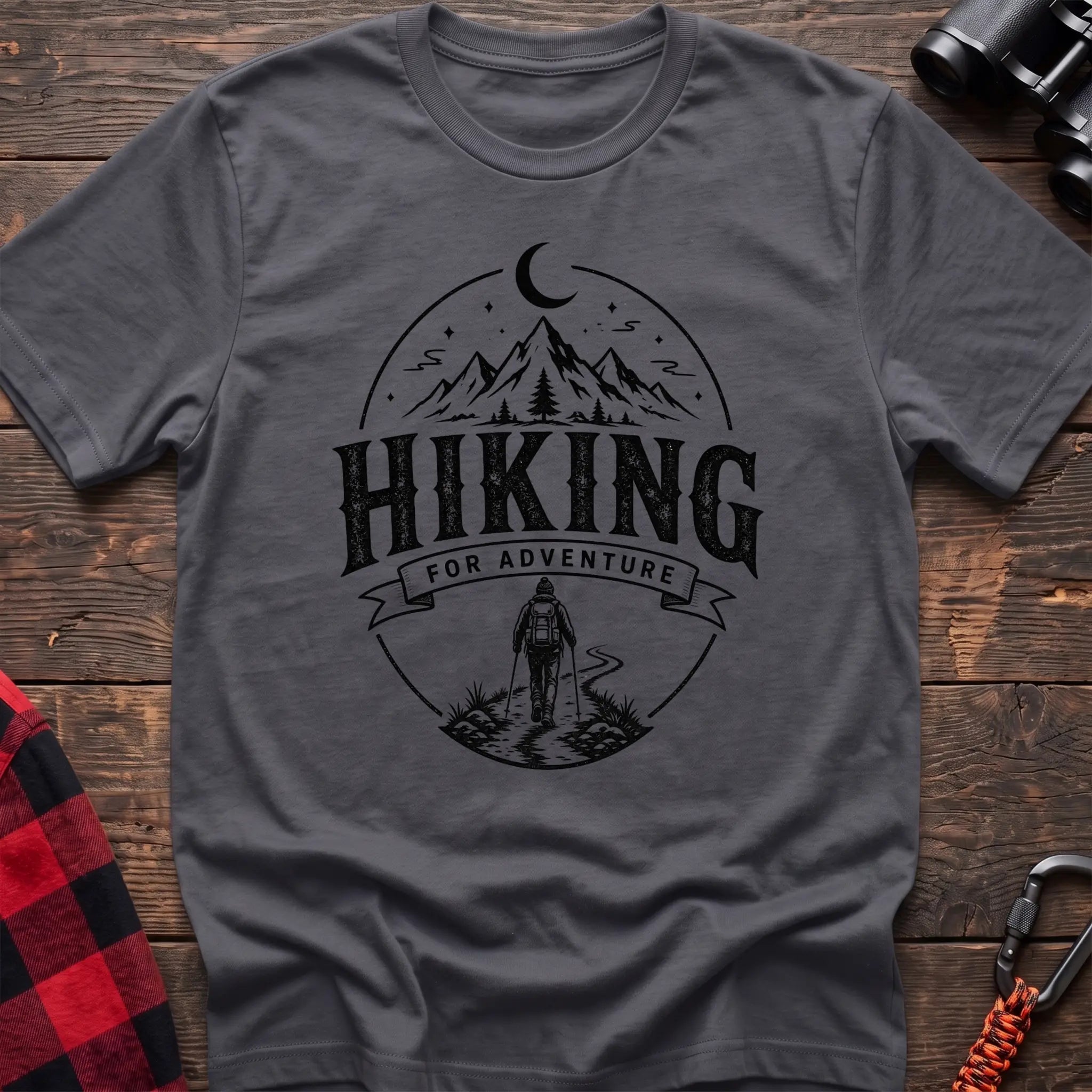 Charcoal cotton t-shirt with vintage Hiking for Adventure graphic of mountains under a crescent moon and a hiker on a trail, flat lay on rustic wood with hiking gear