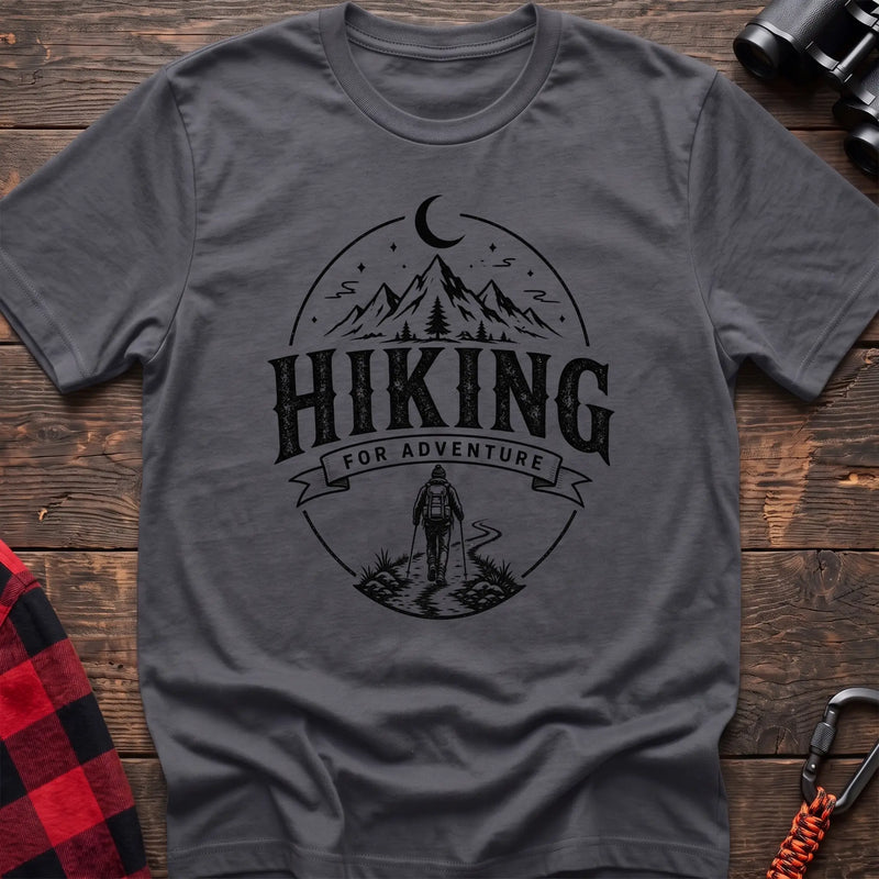 Charcoal cotton t-shirt with vintage Hiking for Adventure graphic of mountains under a crescent moon and a hiker on a trail, flat lay on rustic wood with hiking gear