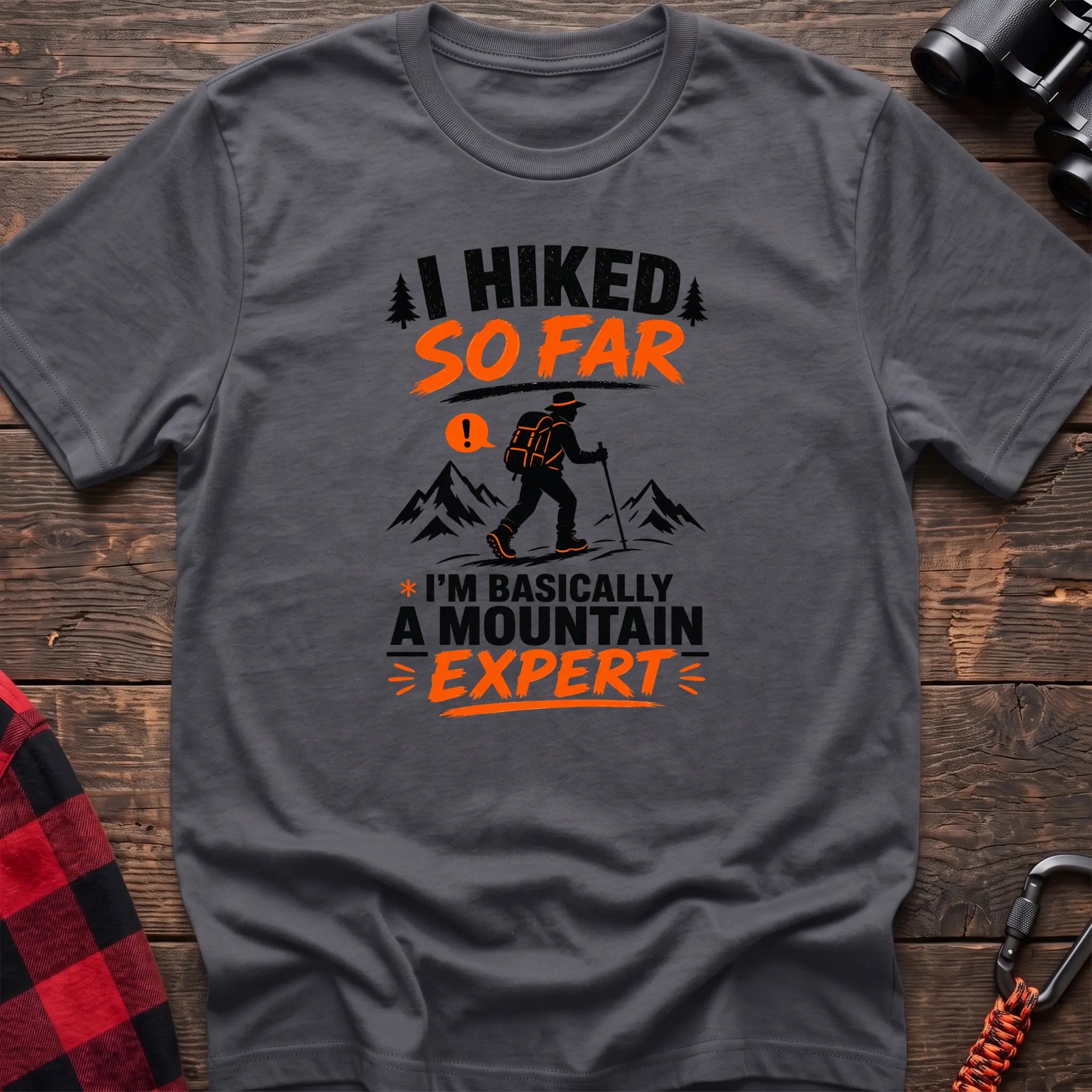 Charcoal cotton t-shirt with I Hiked So Far I'm Basically a Mountain Expert graphic of a hiker with backpack and trekking pole, flat lay on rustic wood with hiking gear