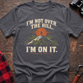 Charcoal cotton t-shirt with a hiker on a summit graphic and I’m Not Over the Hill I’m On It text on rustic wood