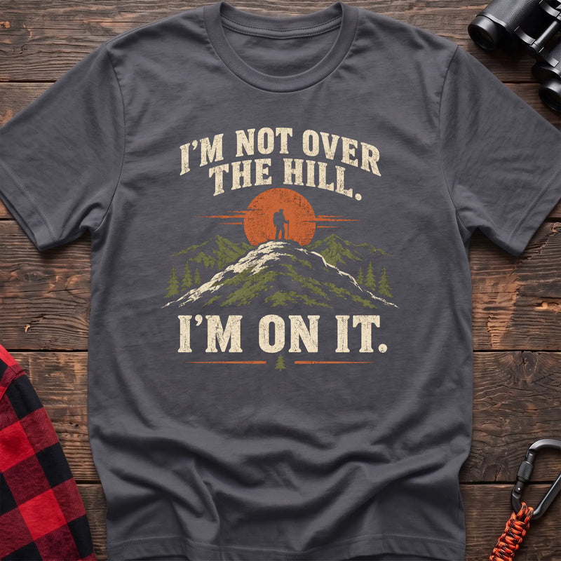 Charcoal cotton t-shirt with a hiker on a summit graphic and I’m Not Over the Hill I’m On It text on rustic wood