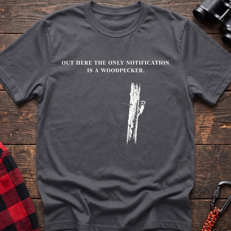 Charcoal cotton t-shirt with a woodpecker graphic and Out Here the Only Notification Is a Woodpecker text on rustic wood