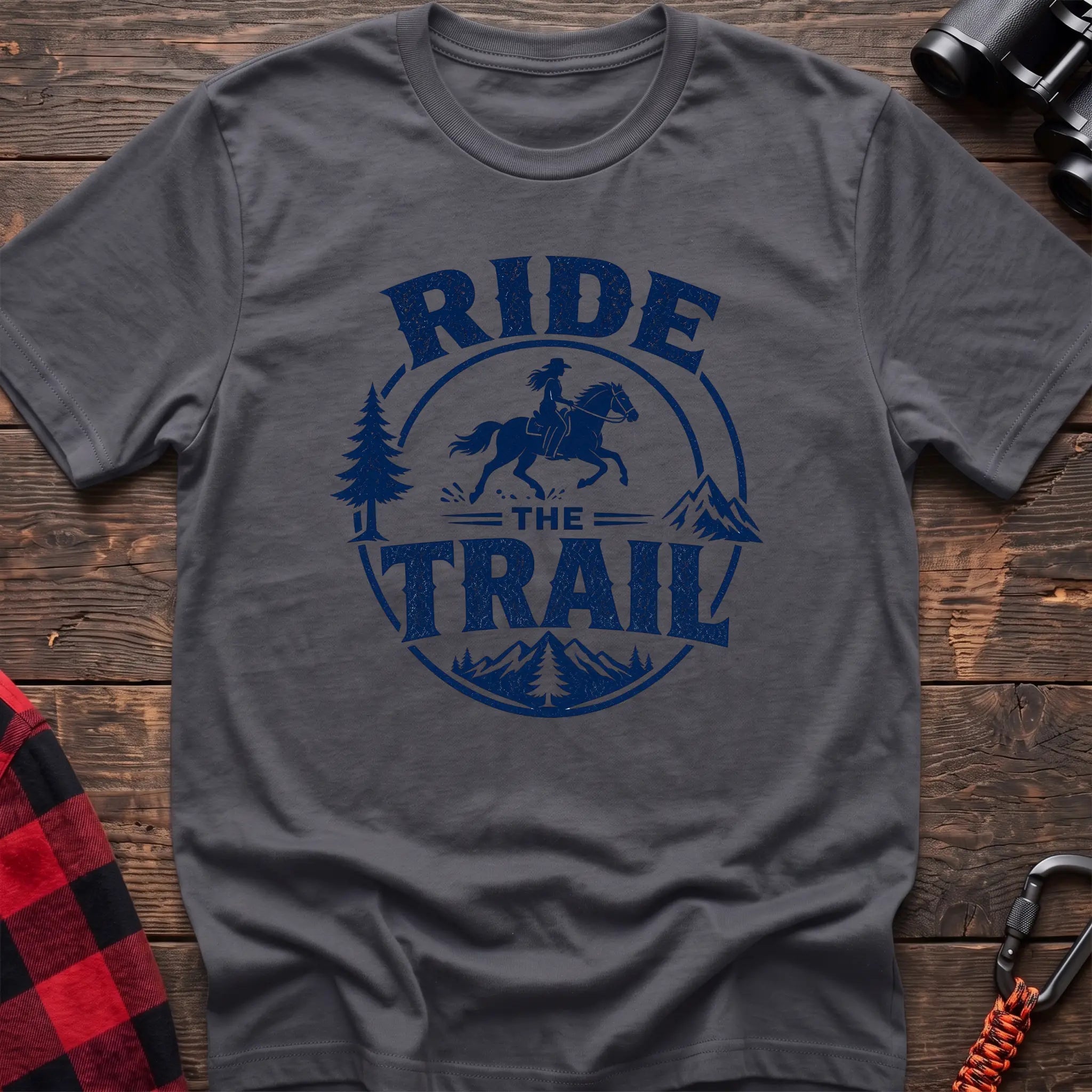 Charcoal cotton t-shirt with navy Ride the Trail graphic of a woman horseback riding through mountains and pine trees, flat lay on rustic wood with hiking gear