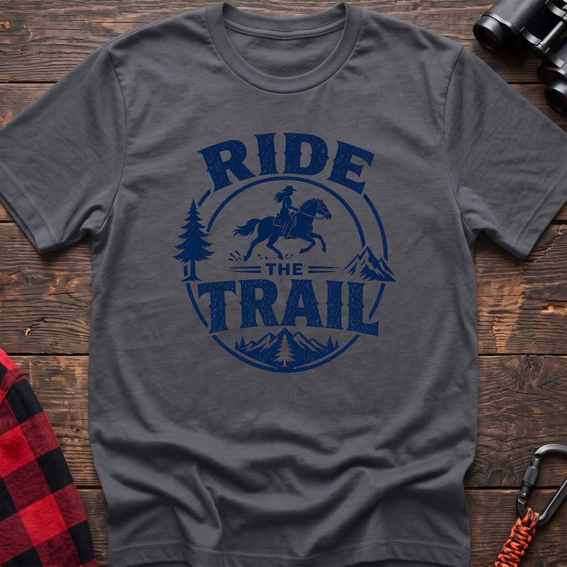 Charcoal cotton t-shirt with navy Ride the Trail graphic of a woman horseback riding through mountains and pine trees, flat lay on rustic wood with hiking gear