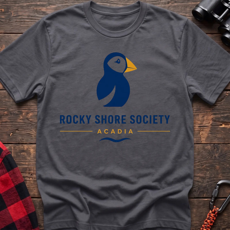 Charcoal cotton t-shirt with a navy puffin logo, yellow beak, and Rocky Shore Society Acadia text on a rustic wood background