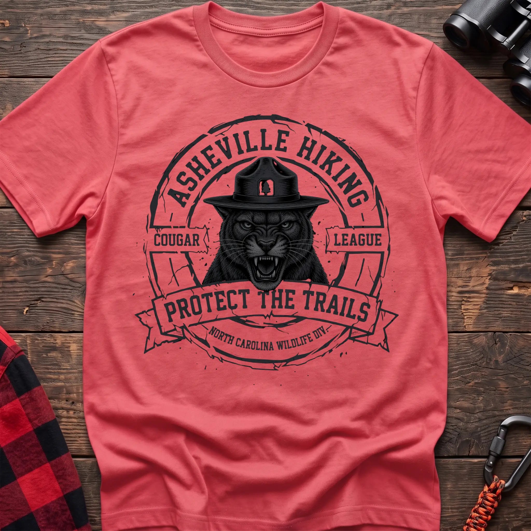 Coral silk cotton t-shirt with vintage Asheville Hiking Cougar League graphic of a roaring cougar in a park ranger hat, Protect the Trails North Carolina Wildlife Division badge, flat lay on rustic wood with hiking gear