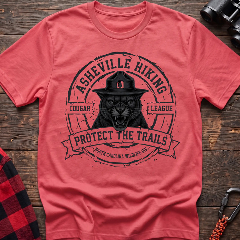 Coral silk cotton t-shirt with vintage Asheville Hiking Cougar League graphic of a roaring cougar in a park ranger hat, Protect the Trails North Carolina Wildlife Division badge, flat lay on rustic wood with hiking gear