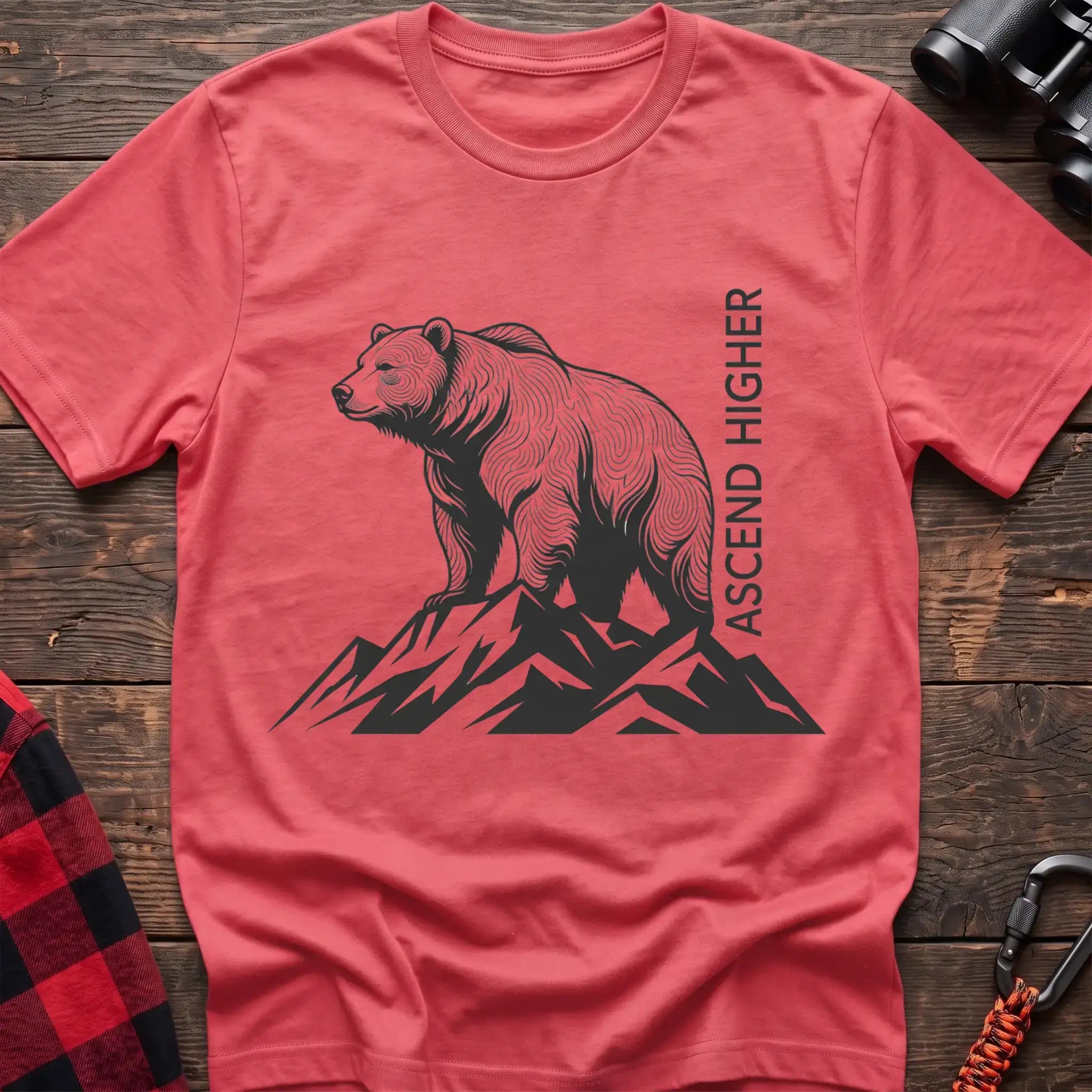 Coral silk t-shirt with a black illustration of a bear standing on mountain peaks, with the vertical text “ASCEND HIGHER” beside it, laid flat on a wooden surface.