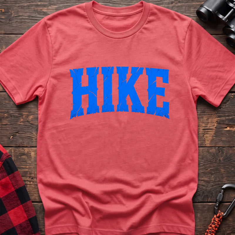 Coral silk cotton t-shirt with distressed arched blue HIKE graphic in cracked varsity lettering, flat lay on rustic wood with hiking gear