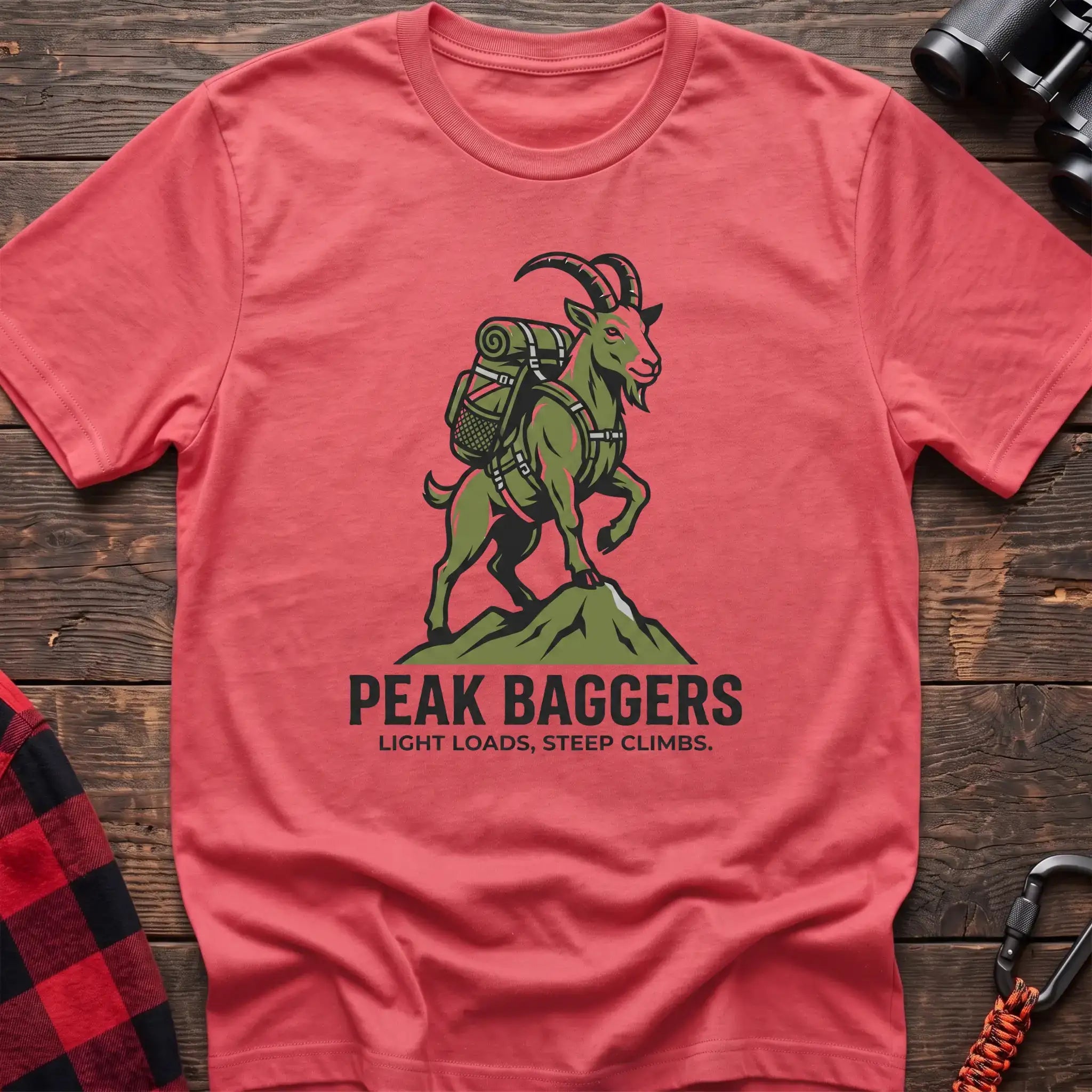 Coral silk t-shirt with an illustration of a mountain goat wearing a backpack standing on a peak, with the text “PEAK BAGGERS” and “LIGHT LOADS, STEEP CLIMBS,” laid flat on a wooden surface.