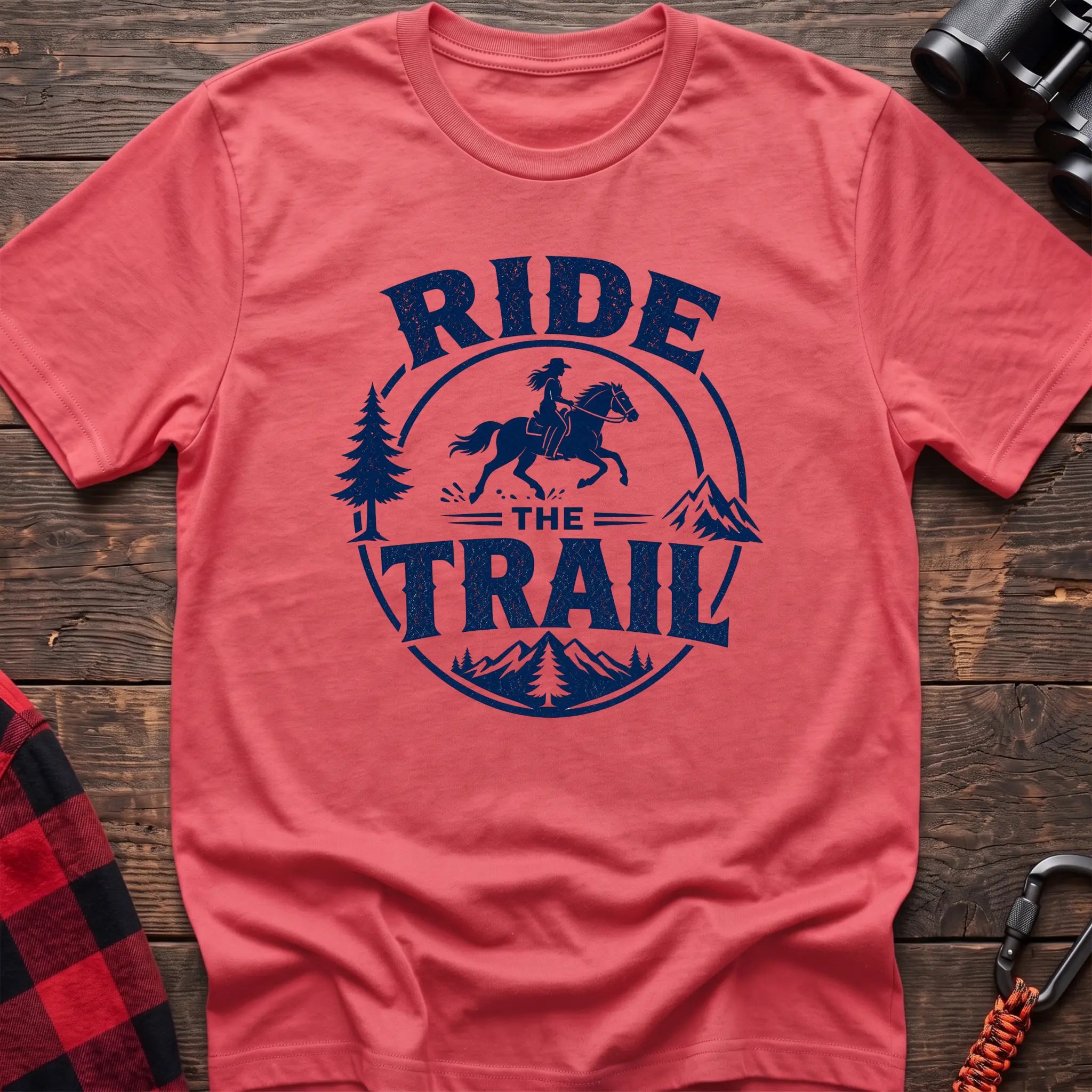 Coral silk cotton t-shirt with navy Ride the Trail graphic of a woman horseback riding through mountains and pine trees, flat lay on rustic wood with hiking gear