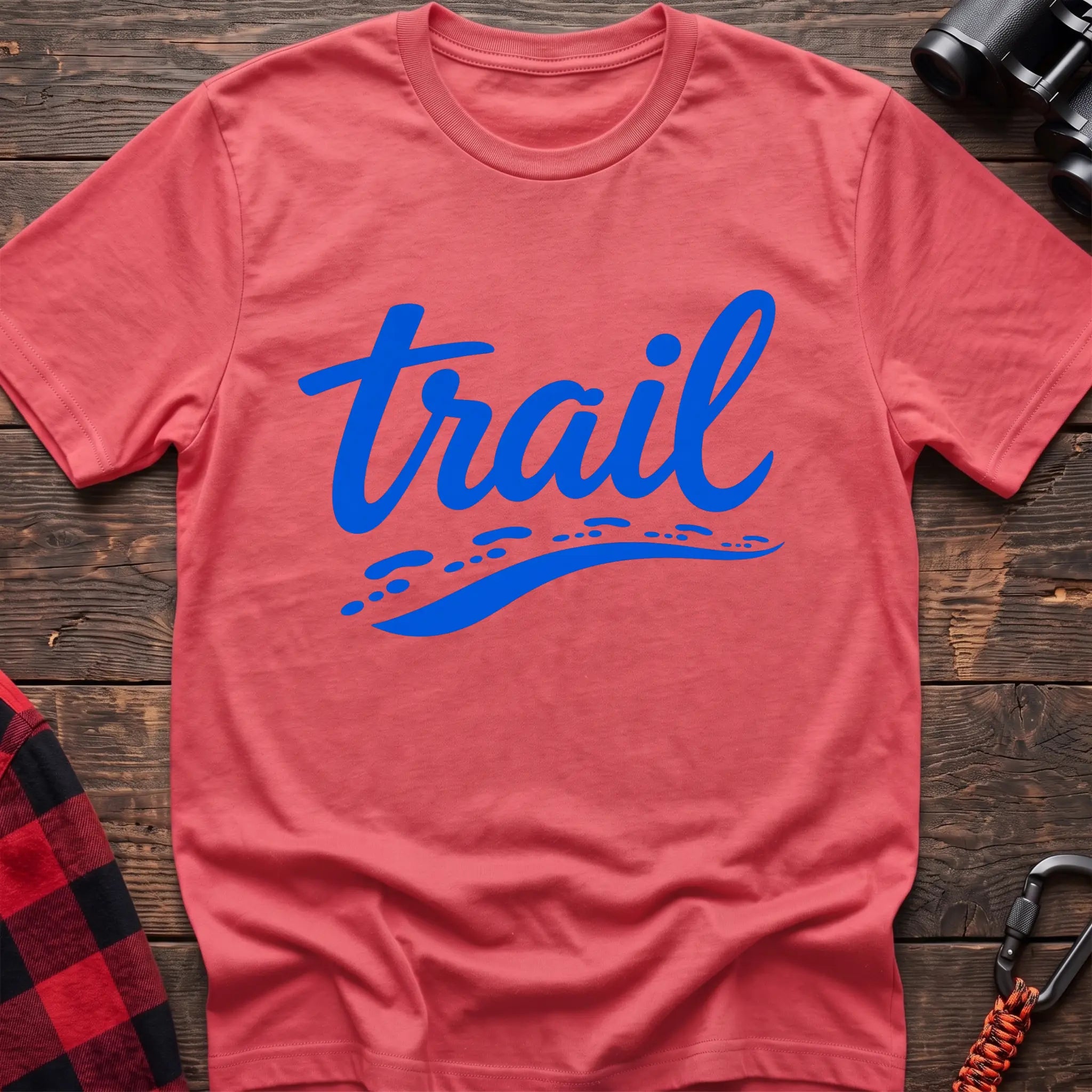 Coral silk cotton t-shirt with blue cursive trail script and dotted path graphic, flat lay on rustic wood with hiking gear