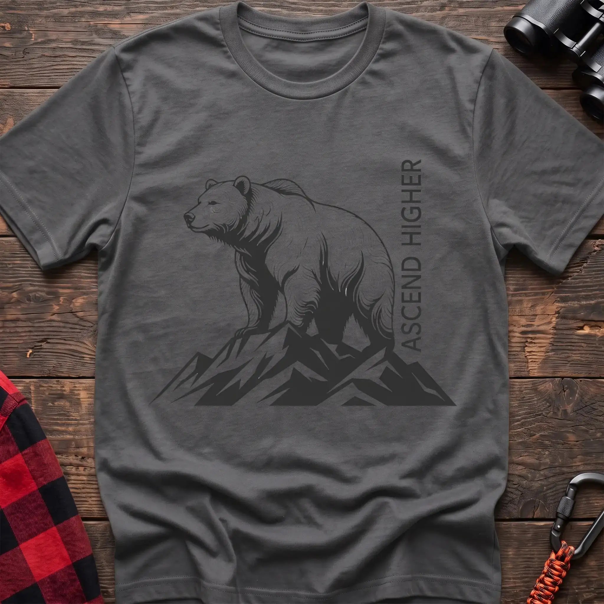 Graphite heather t-shirt with a black illustration of a bear standing on mountain peaks, with the vertical text “ASCEND HIGHER” beside it, laid flat on a wooden surface.