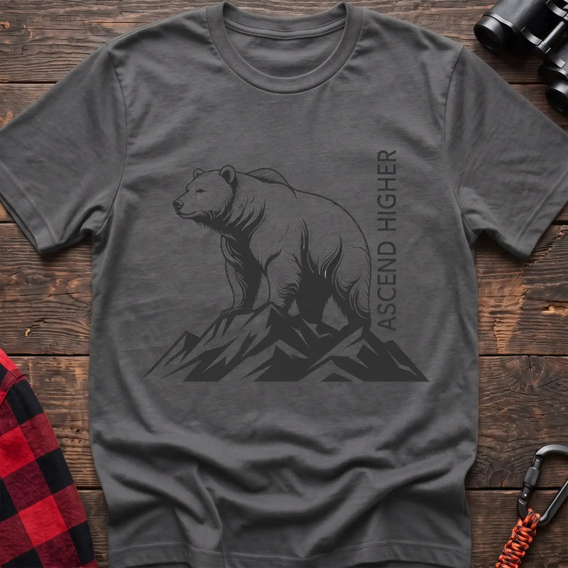 Graphite heather t-shirt with a black illustration of a bear standing on mountain peaks, with the vertical text “ASCEND HIGHER” beside it, laid flat on a wooden surface.
