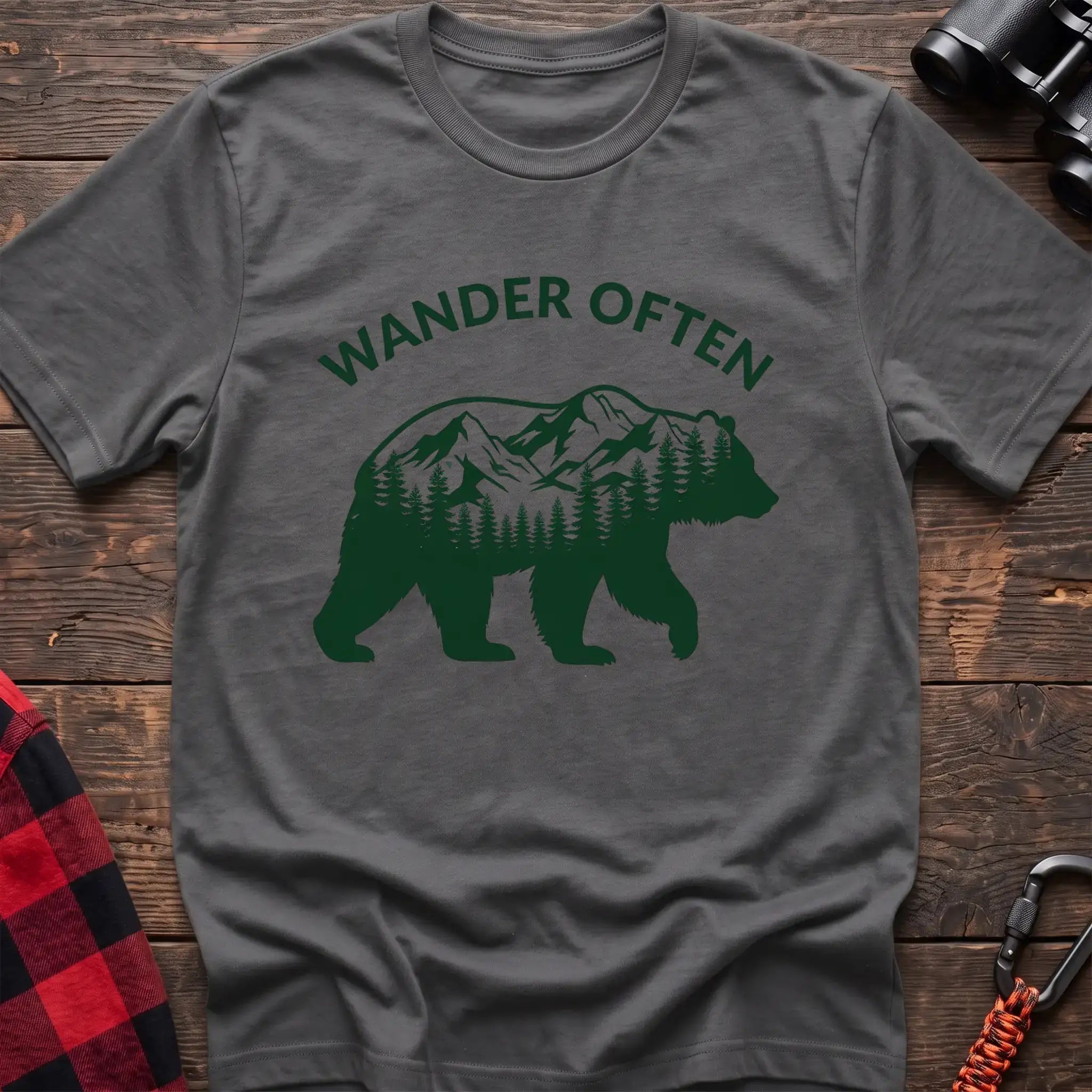 Graphite heather t-shirt with a dark green silhouette of a walking bear containing a mountain and pine forest scene inside, with the arched text “WANDER OFTEN” above it, laid flat on a wooden surface.