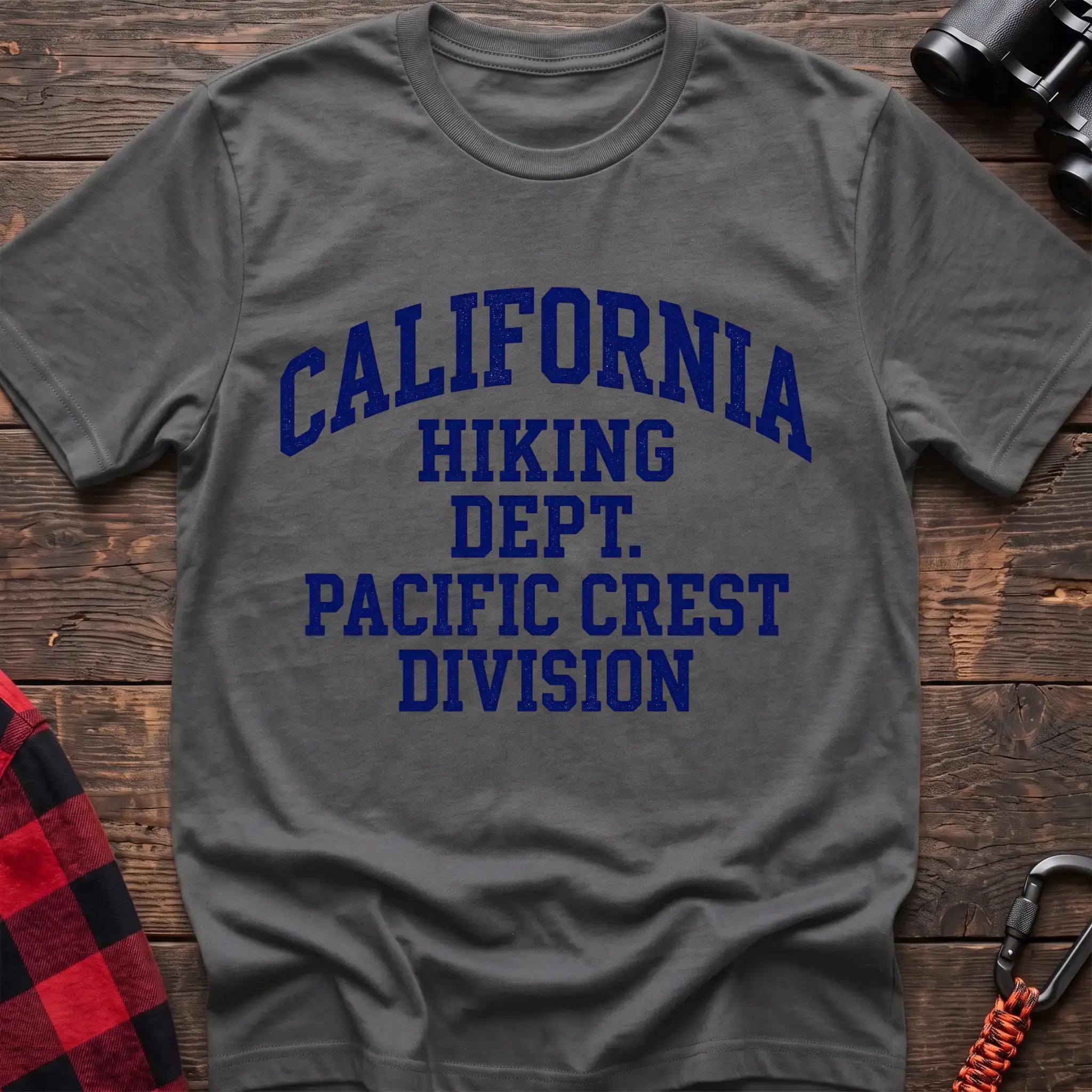 Graphite heather t-shirt with distressed blue collegiate-style text reading “CALIFORNIA HIKING DEPT. PACIFIC CREST DIVISION,” laid flat on a wooden surface.