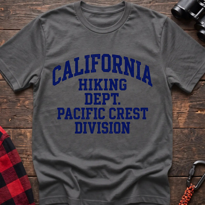 Graphite heather t-shirt with distressed blue collegiate-style text reading “CALIFORNIA HIKING DEPT. PACIFIC CREST DIVISION,” laid flat on a wooden surface.