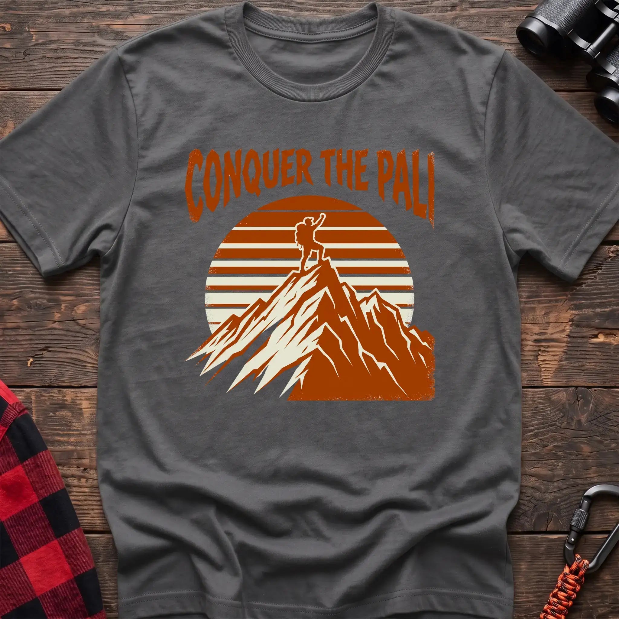Graphite heather t-shirt with orange text reading Conquer the Pali above a hiker standing on a mountain peak with a retro sun graphic