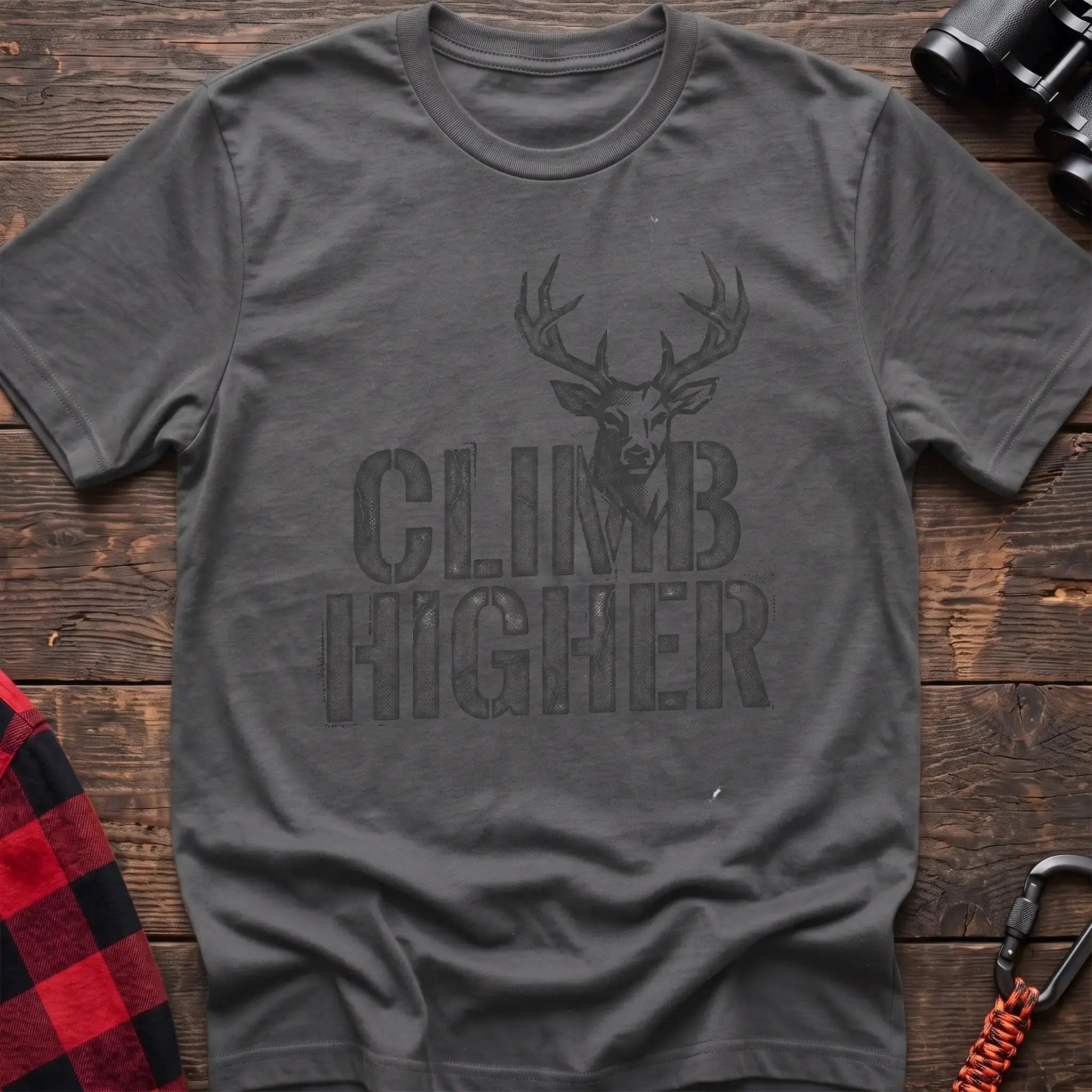 Graphite heather t-shirt with a distressed dark graphic of a deer’s head and antlers rising from the bold text “CLIMB HIGHER,” laid flat on a wooden surface.